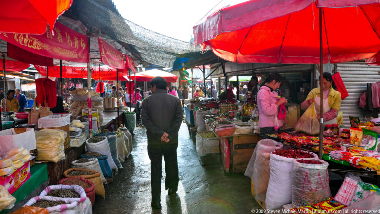 Person walking down a narrow, wet food-market aisle in Lijiang, China, lined with open sacks, bowls, and produce under red umbrellas and tarps.