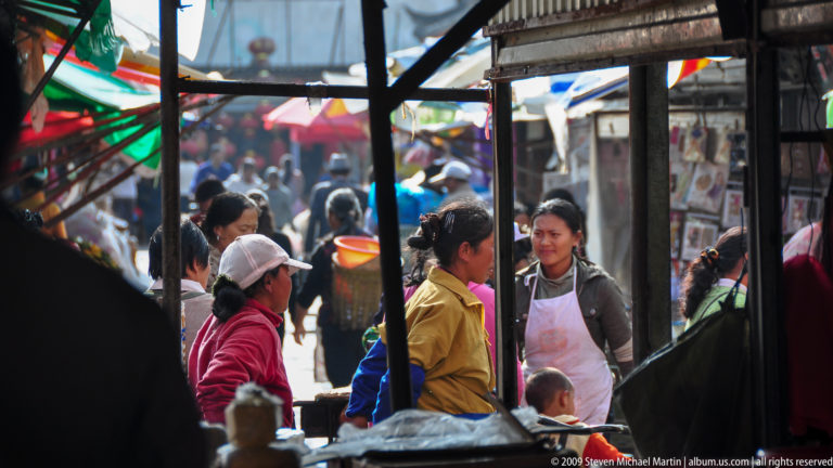 Women vendors talking among crowded stalls in a busy market aisle in Lijiang, Yunnan, China, framed by awning poles and hanging equipment.
