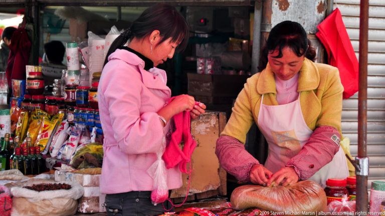 Vendor in a Lijiang market ties a large clear bag over a round bundle on the counter while a customer watches, with shelves of jars and packaged goods behind them.