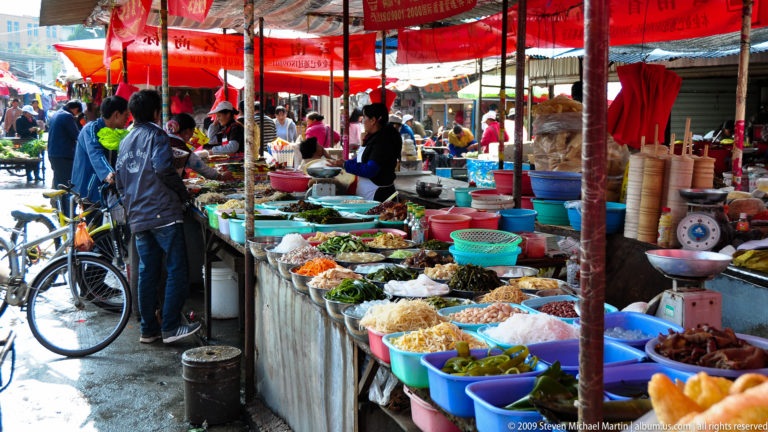 Long row of covered market stalls in Lijiang, Yunnan, with vegetables and prepared foods in colorful tubs and baskets and shoppers under red awnings.