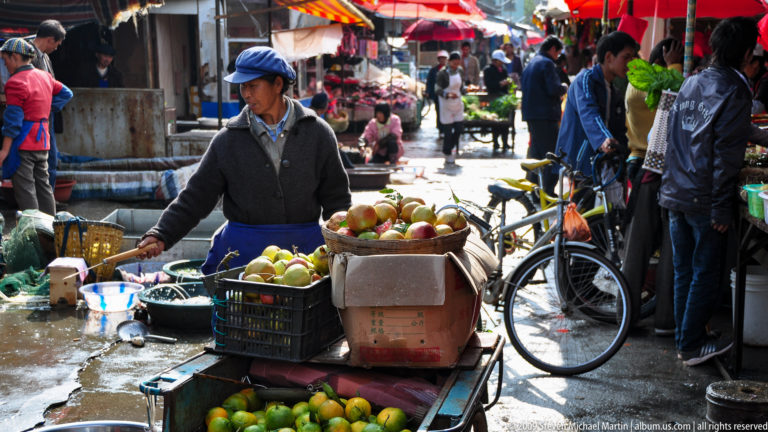 Apples in a large basket stacked above crates of green citrus on a street fruit cart, with a vendor and bicycle beside the stall in Lijiang, Yunnan, China.