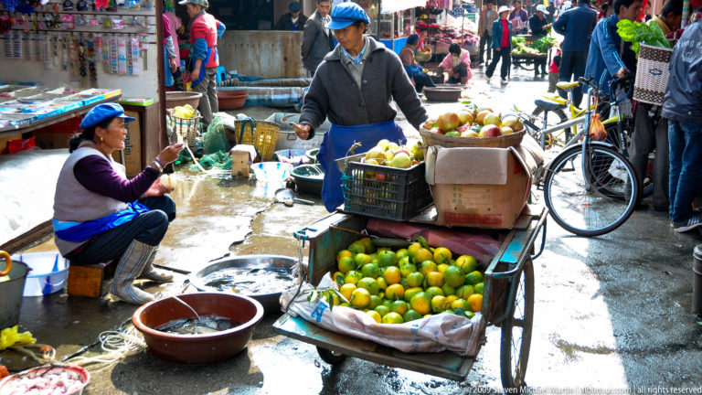 Woman selling green citrus and pale apples from a handcart in a wet market lane in Lijiang, Yunnan, China.