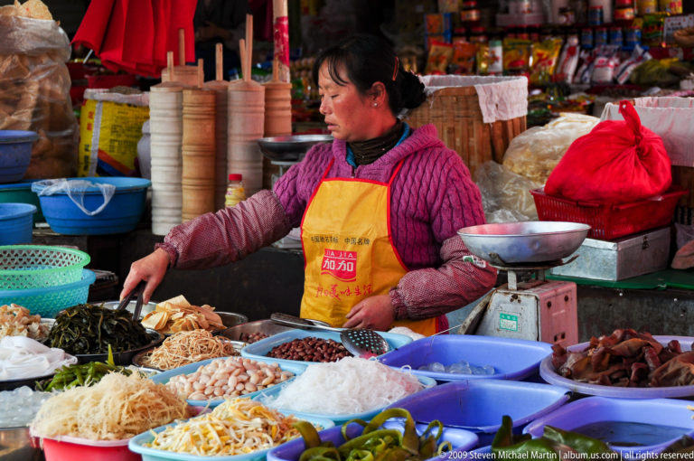 Woman in a Lijiang market standing behind many bowls and tubs filled with noodles, shredded vegetables, beans, and dark preserved foods.