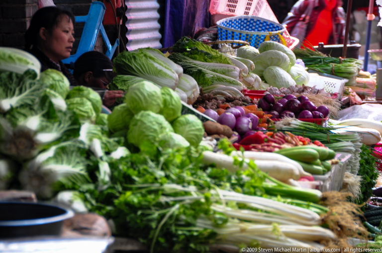 Close-up of neatly arranged vegetables, with leafy greens and pale cabbages in the foreground and eggplants, peppers, and root vegetables receding into the distance at a market in Lijiang, Yunnan, China.