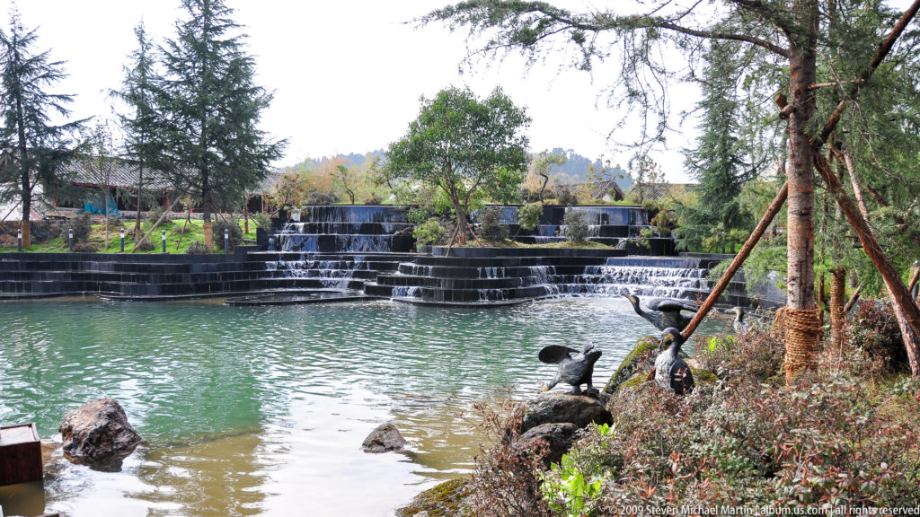 Terraced artificial waterfall in a Lijiang garden with water flowing over broad dark stone steps into a pond surrounded by trees and planted banks.