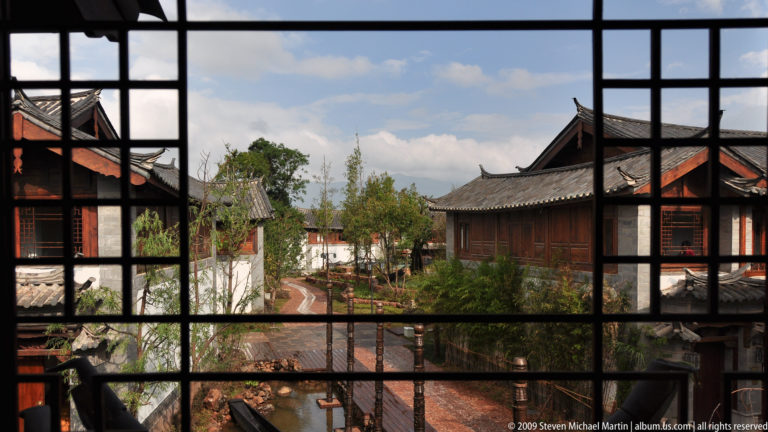 View through a black wooden lattice window onto a traditional Lijiang courtyard with tiled roofs, paths, trees, and a small pond.
