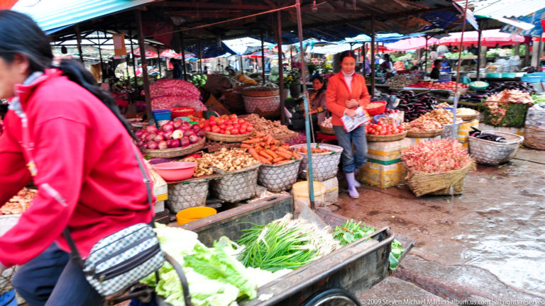 Woman standing behind a vendor stall at an outdoor vegetable market in Lijiang, Yunnan, with open baskets of leafy greens, onions, carrots, and potatoes under tarps.