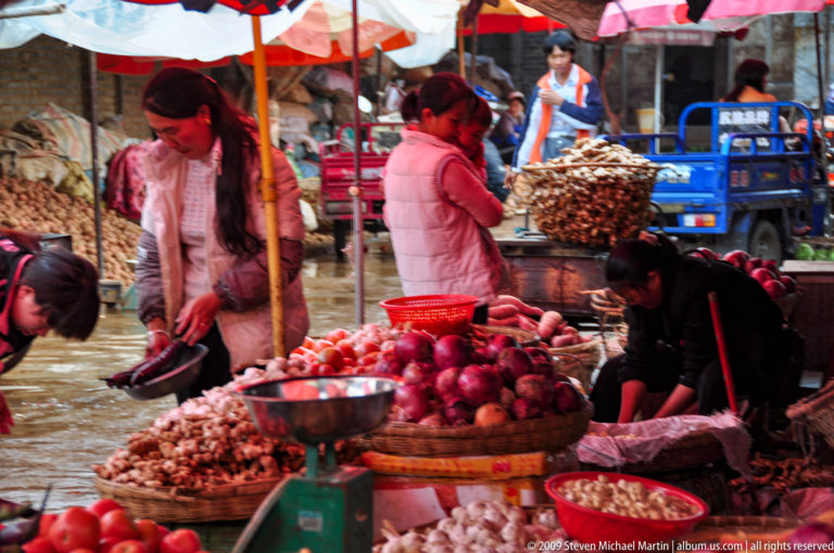 Women selling onions, garlic, ginger, and other produce from heaped baskets of red onions and pale roots under umbrellas on a wet street market in Lijiang, Yunnan, China.