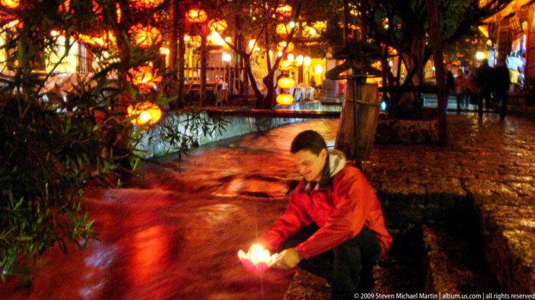 Male visitor crouching by a canal in Lijiang, Yunnan, holding a lit lotus lamp near the water with lantern-lit trees in the background.
