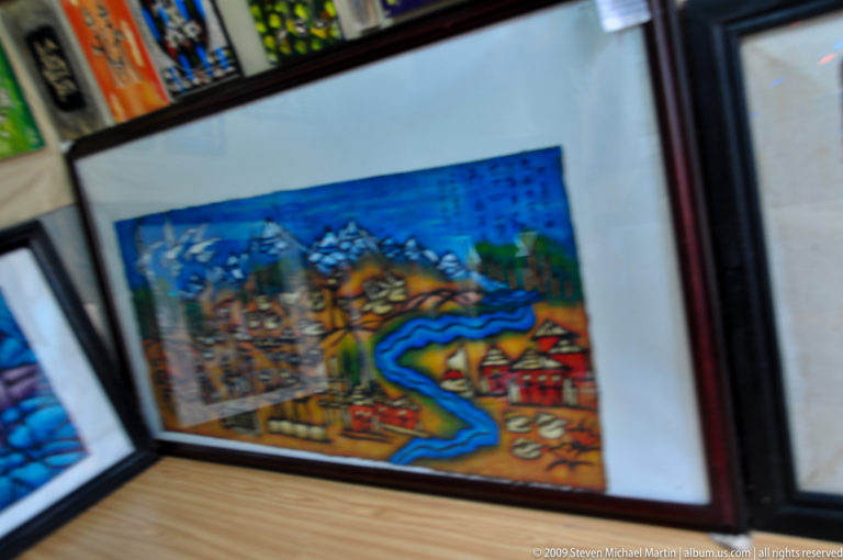 Framed colorful folk artwork on a tabletop showing stylized mountains, a settlement, and a bright blue river.