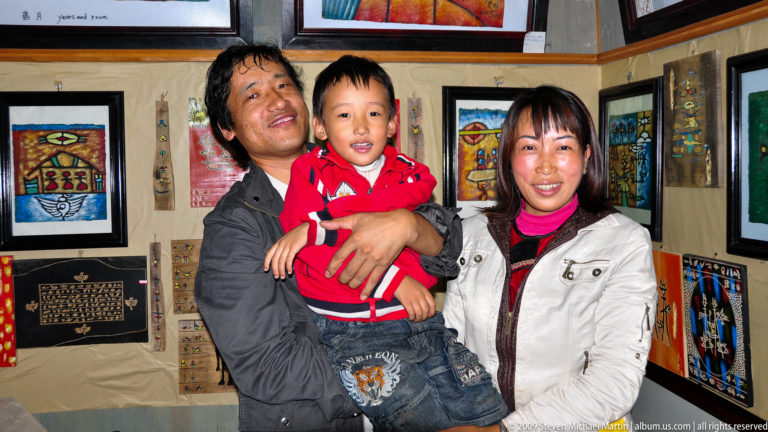 Two adults hold a small child and face the camera inside an art shop with colorful framed artworks on the walls.