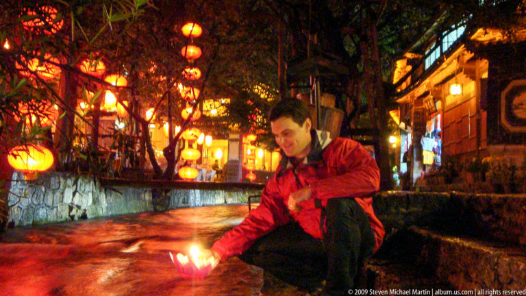 Male visitor crouching by a stone canal at night in Lijiang, holding a glowing paper lotus lamp above the rushing water.