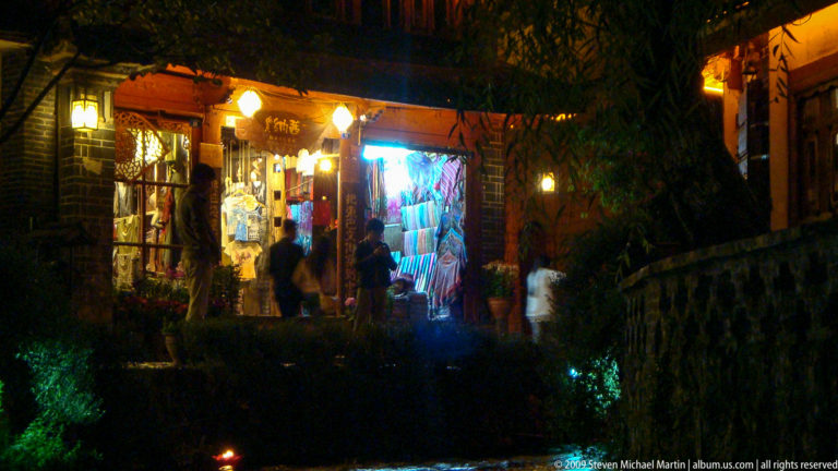 Small illuminated textile or souvenir shop beside a dark canal in Lijiang, Yunnan, China, with brightly lit merchandise filling the doorway above stone edging.