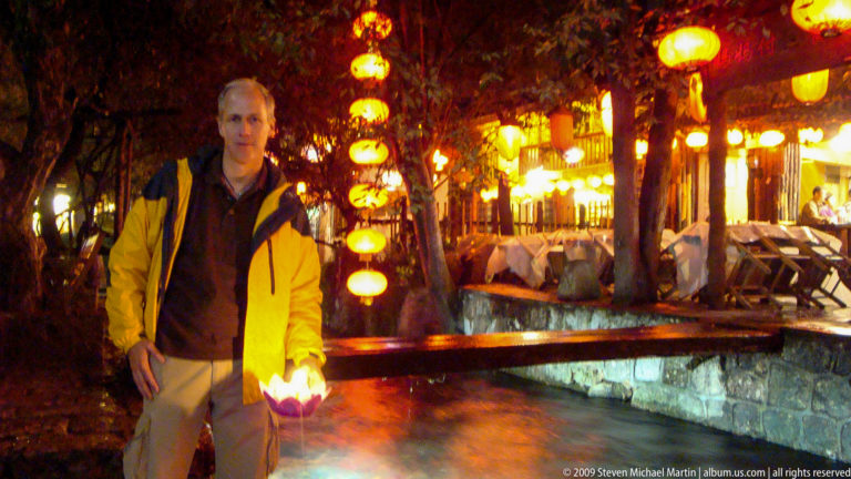 Male visitor holding a glowing lotus paper lamp beside a rushing canal lined with lanterns and restaurant terraces in Lijiang, Yunnan, China.
