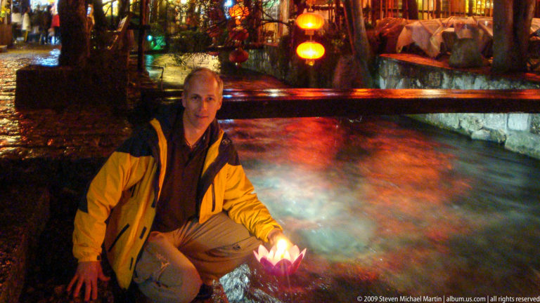 Male visitor crouching at night beside a canal in Lijiang, China, holding an illuminated folded-paper lotus lamp over the water with colorful lantern reflections and a bridge rail in the background.
