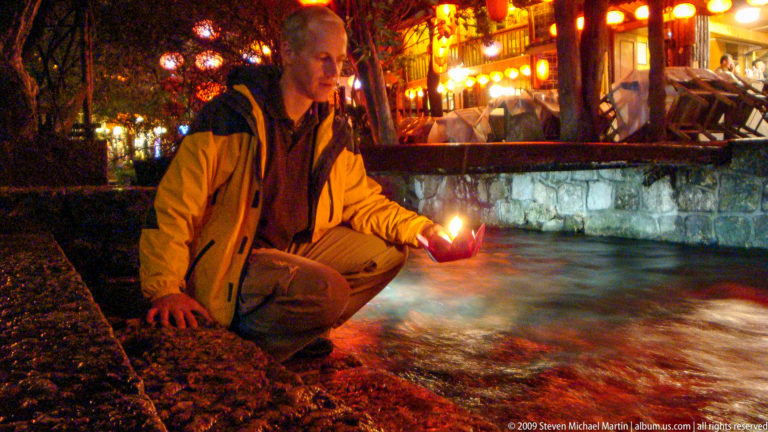 Male visitor crouching beside a fast-moving canal in Lijiang, China, holding a lit paper lotus over the water with lantern-lit terraces and a stone wall in the background.