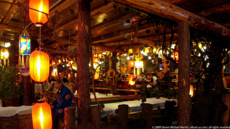 Open-air courtyard restaurant in Lijiang, China, with glowing lanterns, timber galleries, and diners seated around a central water feature.
