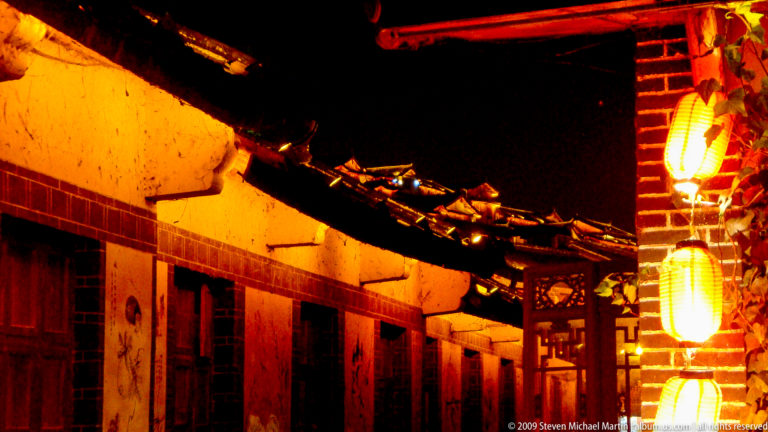 Whitewashed Old Town wall in Lijiang at night, with timber eaves, lanterns, and a curving tiled roofline over dark doors lit by warm orange light.
