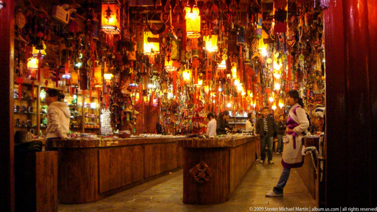 Interior of a large souvenir shop in Lijiang, Yunnan, with hanging crafts, decorative goods, wooden counters forming aisles, and several people under warm lighting.
