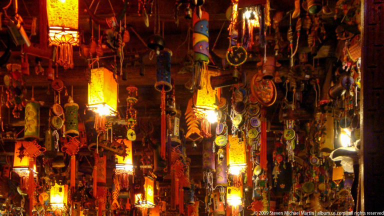 Medium view of a warmly lit souvenir stall in Lijiang, with hundreds of lanterns, red tassels, metal ornaments, and woven trinkets hanging in dense layers.