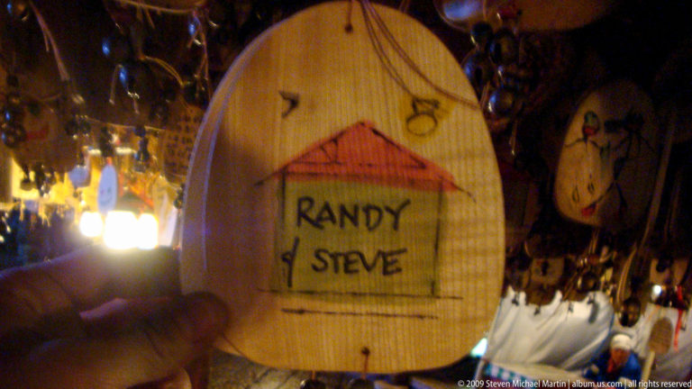 Close-up of a wooden plaque in Lijiang, China, reading “Randy & Steve” in black inside a simple house outline, hanging by a cord among other souvenirs.
