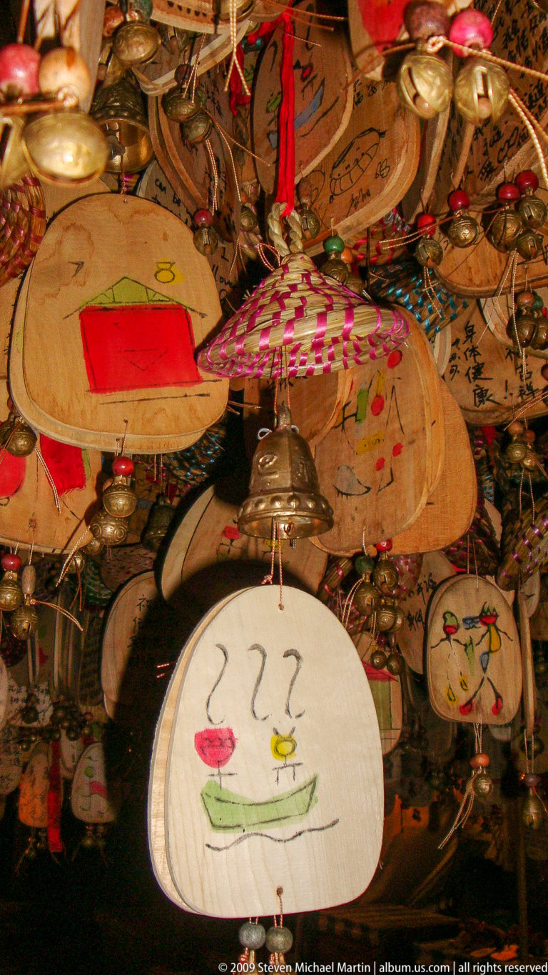 Hanging wooden wish plaques with handwriting and drawings, interspersed with bronze-colored bells on red cords in a Lijiang market in Yunnan, China.