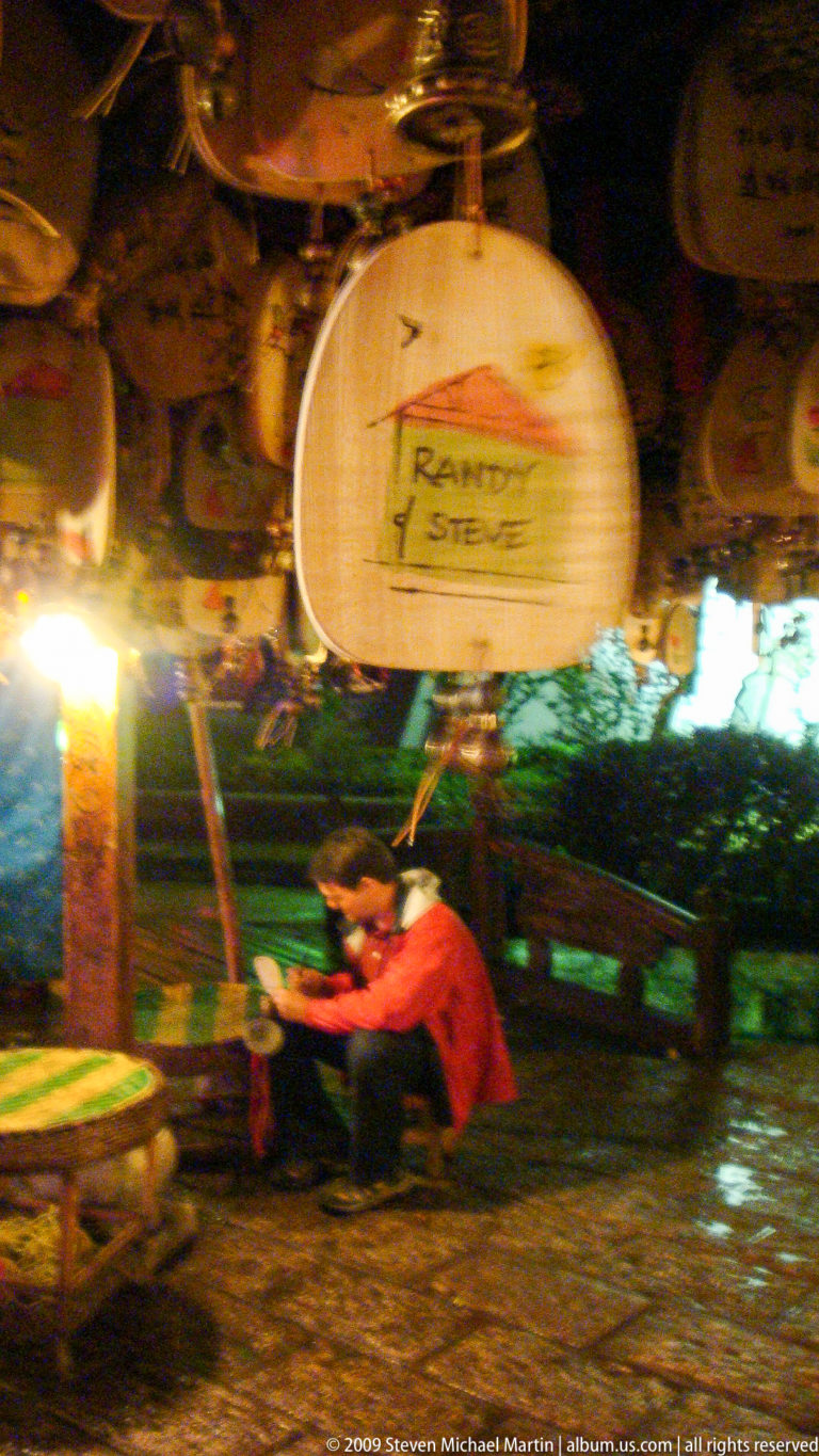 Wooden souvenir plaque in Lijiang, Yunnan, China, with handwritten names “Randy & Steve” in focus and a blurred seated visitor and shop display in the background.
