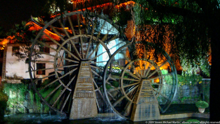 Three traditional wooden waterwheels beside a building with timber flumes and stone-lined water channels in Lijiang, Yunnan, China.