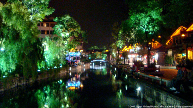 Night view of a tree-lined canal in Lijiang, China, with a low arched bridge and shopfront lights reflected in the dark water.