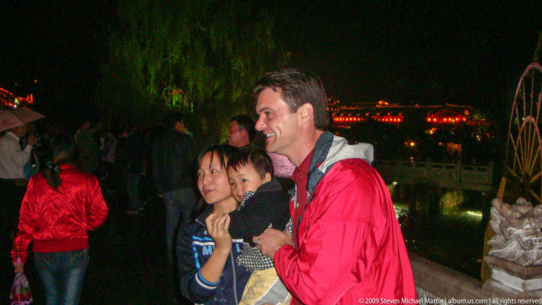 Male visitor posing in profile with a woman holding a small child at night, with illuminated roofs and trees in the background in Lijiang, Yunnan, China.