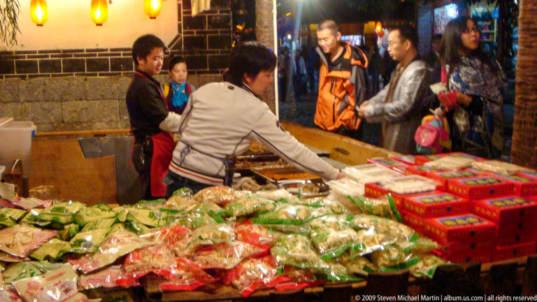 Vendor serving packaged green and pale foods to customers at a night market stall in Lijiang, Yunnan, China.