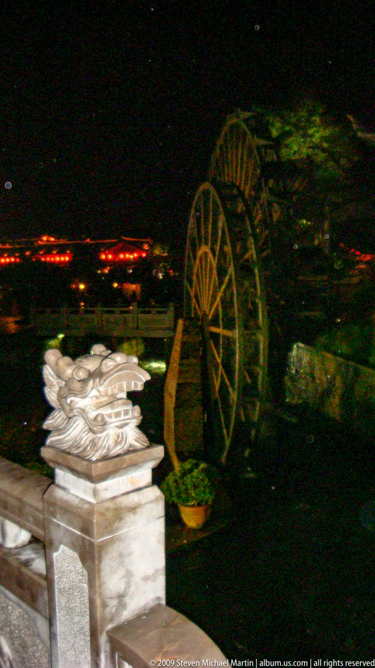 Stone lion or dragon head sculpture in front of a large wooden wheel and lit bridge at night in Lijiang, Yunnan, China.