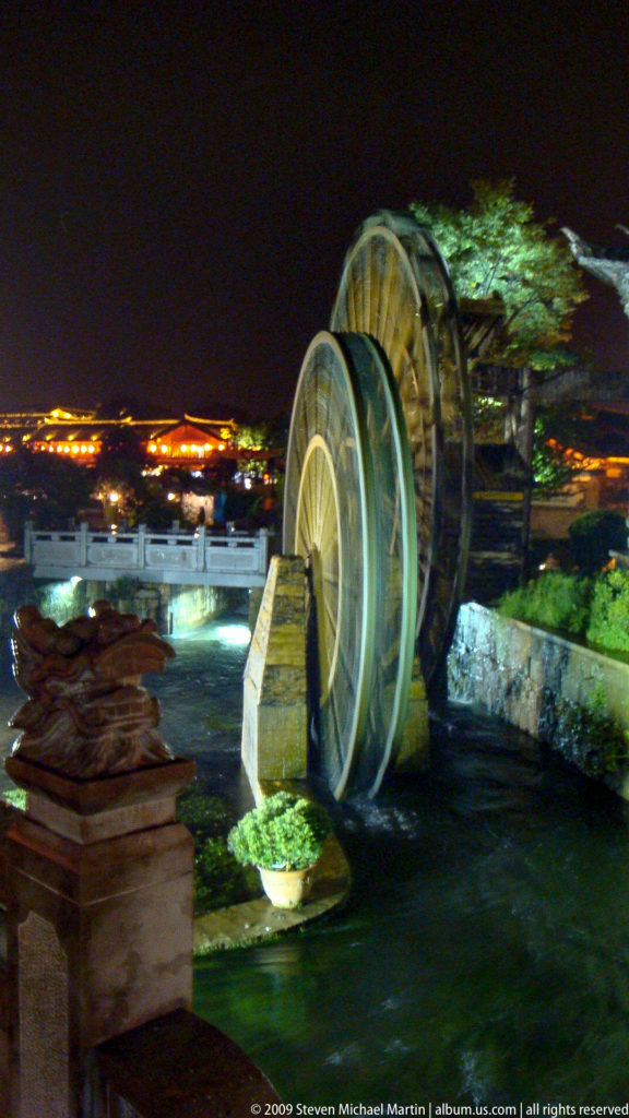 Large wooden waterwheel in a flowing canal next to a stone bridge and carved stone animal head in Lijiang, China.