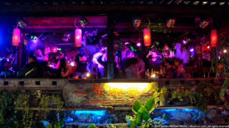 Crowded nightclub in Lijiang, Yunnan, China, with patrons under purple and blue lighting facing a raised stage decorated with red lanterns.