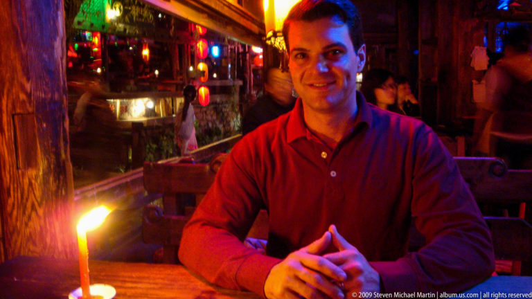 Male visitor in a red shirt sits with hands clasped at a lantern-lit restaurant table in Lijiang, Yunnan, China, with warm lights and other patrons behind him.