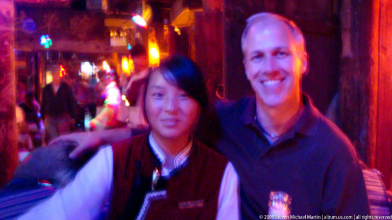 Male visitor and young local woman smiling at the camera in a nightlife venue in Lijiang, Yunnan, China, with colored lighting and other patrons behind them.