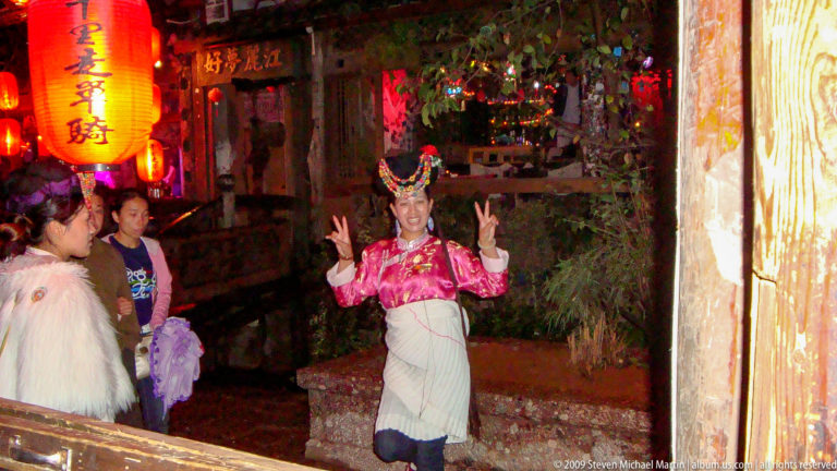 Female performer in bright ethnic-style costume with decorated headdress, pink blouse, and pleated white apron-skirt posing with raised hands while bystanders watch in Lijiang, Yunnan, China.