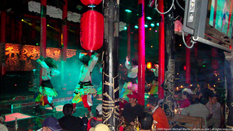Costumed women perform a dance under colored lights in a crowded Lijiang nightlife venue, with audience members seated at tables around red columns.