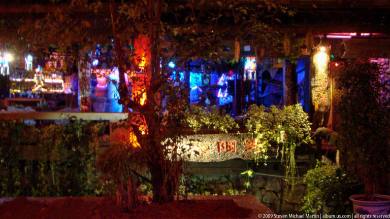 Night view of a garden-fronted bar or performance venue in Lijiang, Yunnan, China, with blue and red stage lighting shining through foliage above stone terrace walls and planted beds.