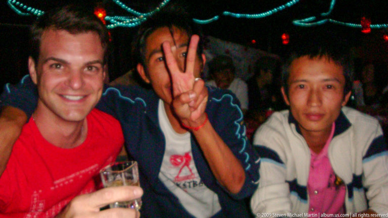 Three men sit at a table in a dark Lijiang venue, one giving a peace sign and another holding a drink, with colored string lights and other patrons in the background.