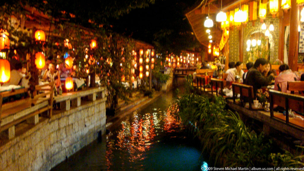 Night view of an illuminated canal in Lijiang Old Town, with red and yellow lanterns reflected in the narrow watercourse and stone balustrades and restaurant terraces on both sides.