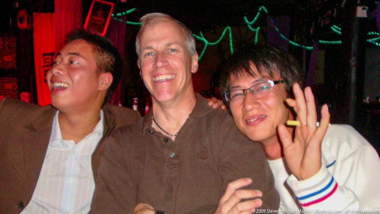 Three men pose together in a club or bar in Lijiang, with the central man smiling as his companions lean in and green rope lights glow in the dark background.