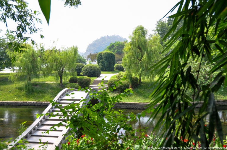 White arched footbridge over a narrow canal in a formal garden with trimmed shrubs, willow trees, and a mountain silhouette in the background in Lijiang, Yunnan, China.