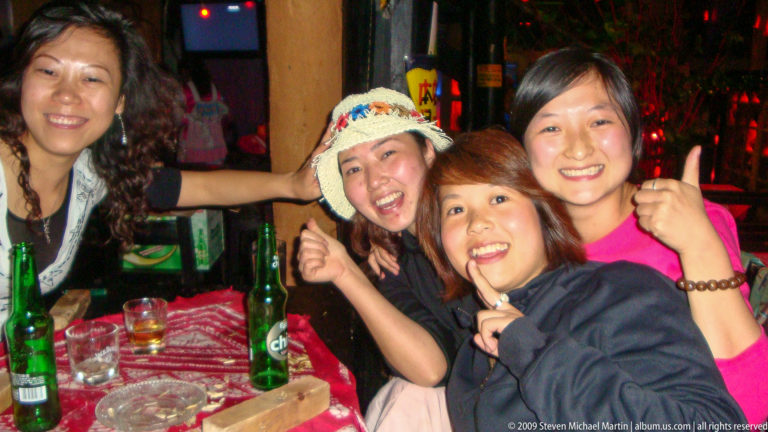 Four young women smiling and giving thumbs-up gestures beside bottles and glasses under dim red and green club lighting.