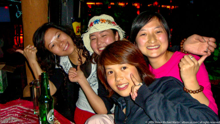 Four young women smiling and leaning together at a table with beer bottles on a red patterned table covering in Lijiang, Yunnan, China.