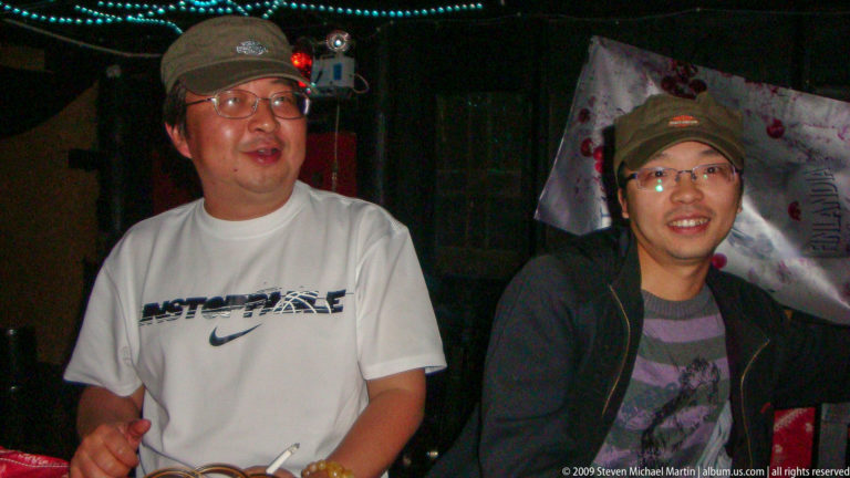 Two men wearing casual shirts and glasses sit facing the camera in a dark entertainment venue with colored lighting and wall décor in the background.
