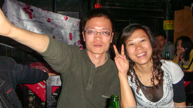 Young man and woman posing together at a table with bottles and dim colored nightlife lights in Lijiang, China; the woman smiles and makes a peace sign.