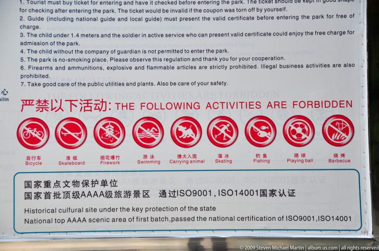 Bilingual park sign in Lijiang, Yunnan, China, showing red pictograms banning bicycles, skateboards, fireworks, swimming, pets, skating, fishing, ball games, and barbecue, and English text noting a national top AAAA scenic area and cultural-site protection unit.