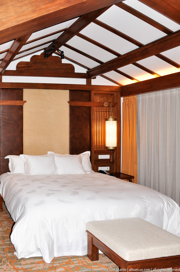 Hotel guest room in Lijiang, China, with a large bed with white linens, timber headboard, wood beams, pale curtains, and a cylindrical lantern-style light.
