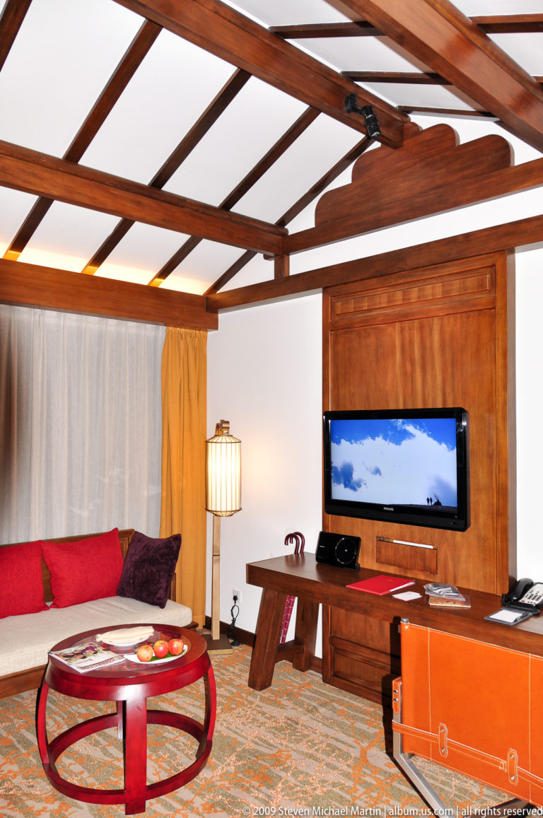 Hotel room interior in Lijiang with a flat-screen TV inset into a timber-paneled wall, red cushions, a lamp, and white plaster walls.